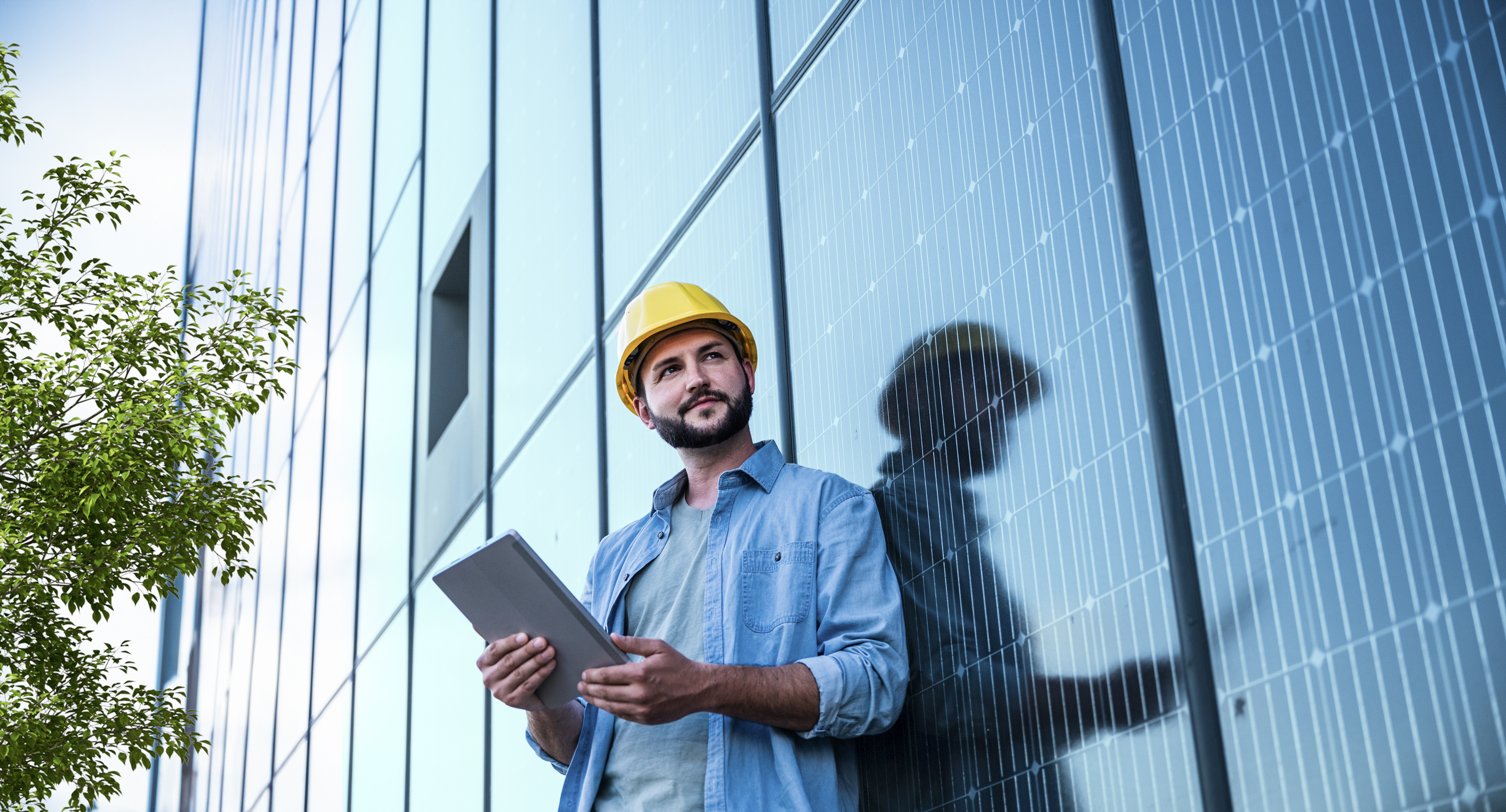Ein Mann in blauem Hemd trägt einen gelben Schutzhelm und lehnt an einer Häuserglasfront