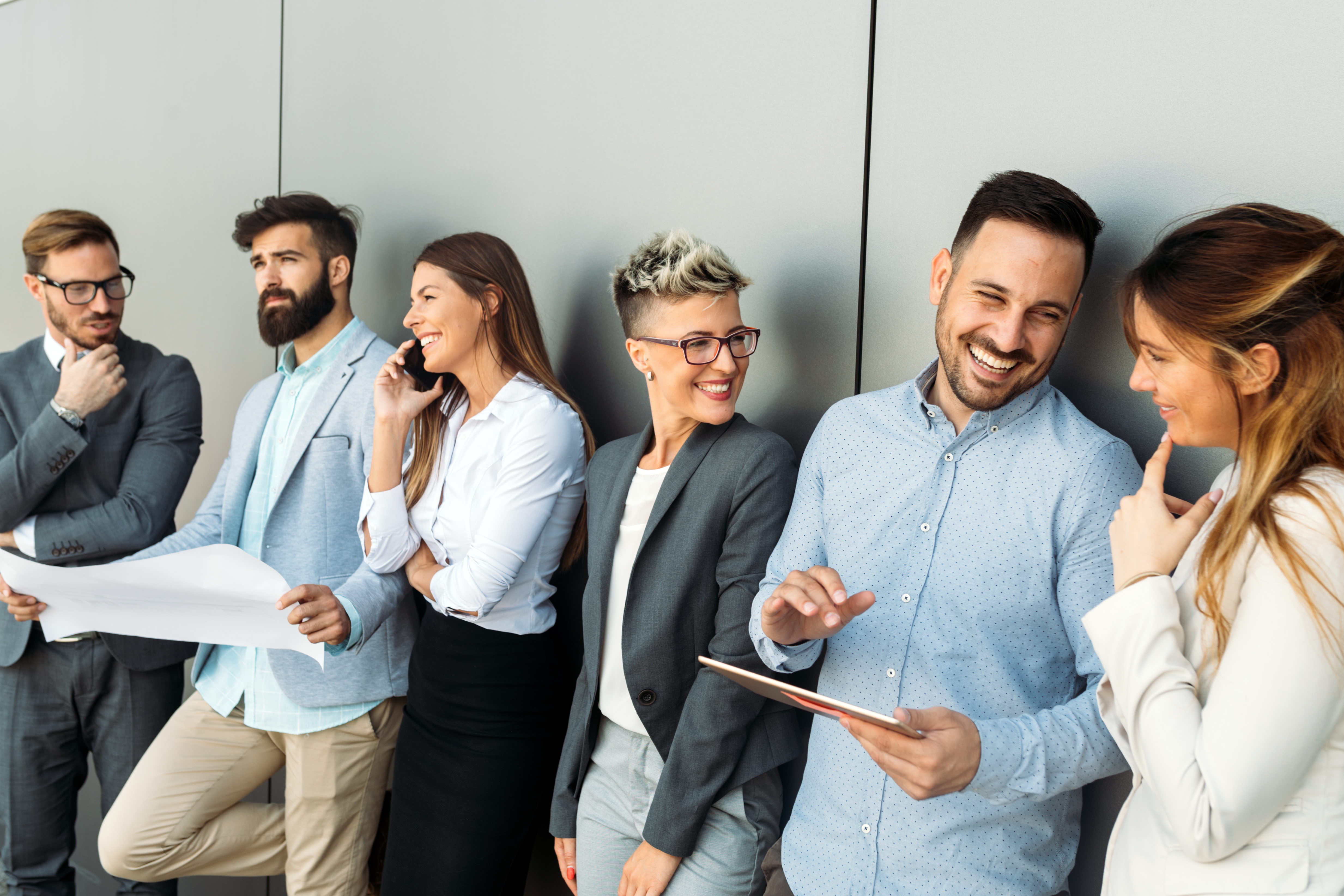 Ein großes Team von Männern und Frauen unterhält sich lachend an eine Bürowand gelehnt